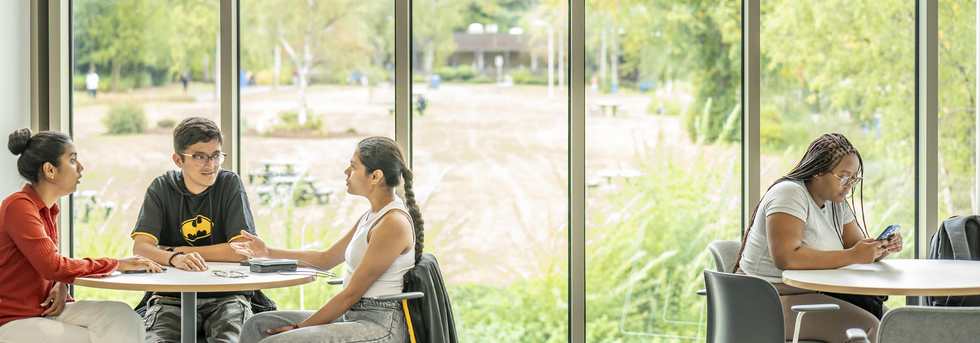Four students sit at two round tables by large windows, three engaged in conversation and one focused on her phone.