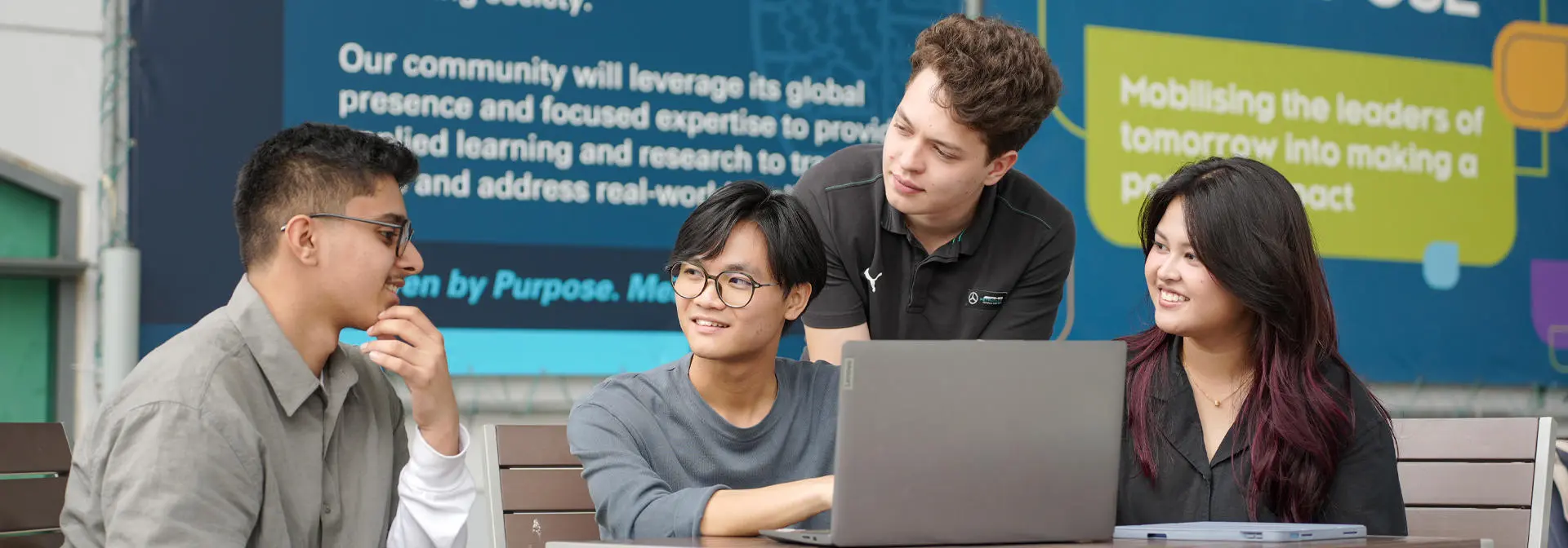 Four students collaboratively working on a laptop at a table, with a colorful wall display behind them.
