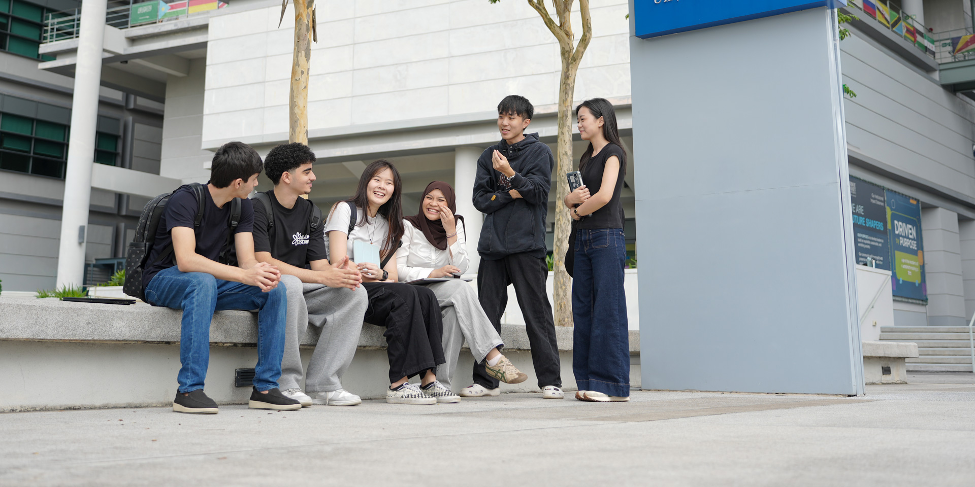A group of students sitting and standing on a bench outdoors, engaged in conversation.