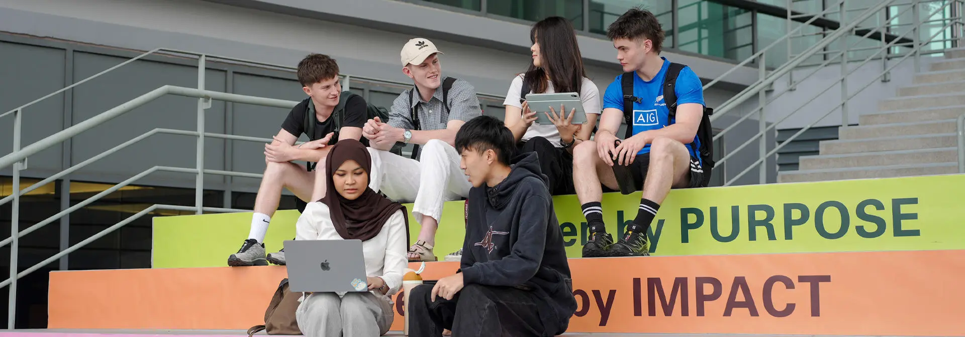Students sit on colorful steps, engaged with laptops and tablets, in a modern outdoor setting.