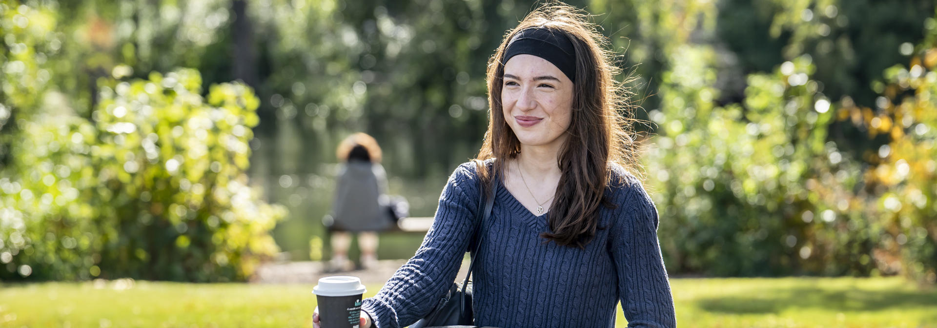A student sits at a wooden table on campus, holding a coffee cup, surrounded by lush greenery.