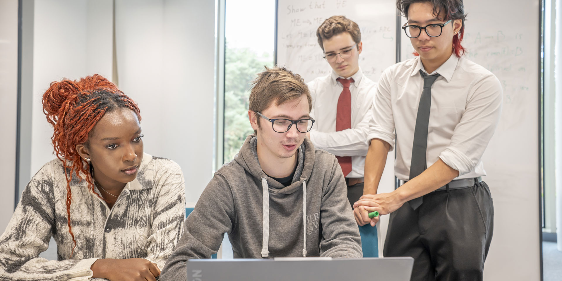 Four students collaborate on a laptop in a modern office setting, focused on their work and sharing ideas.