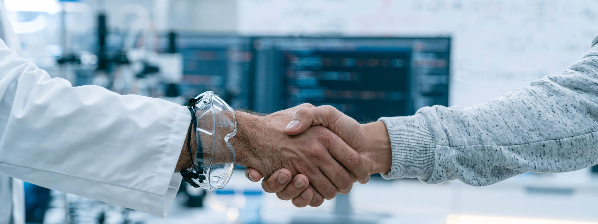 Two hands in a handshake. One with lab goggles and lab coat and another in a jumper.