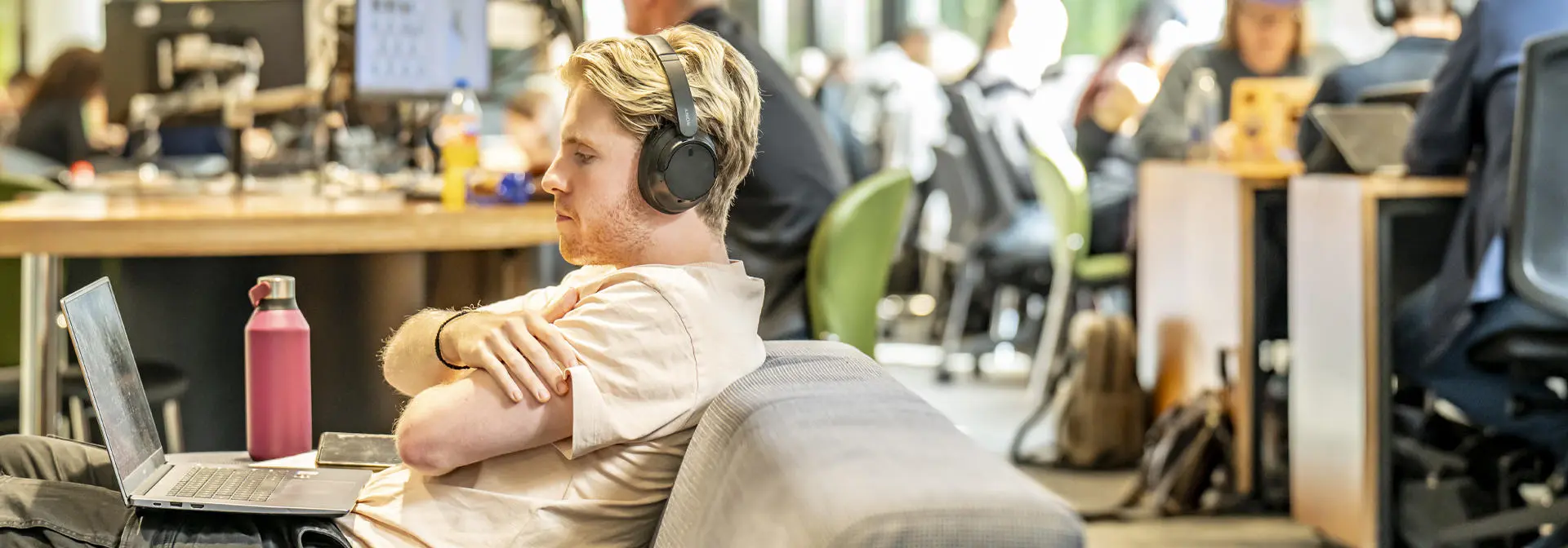 A student seated on a couch, wearing headphones, using a laptop in a busy environment.