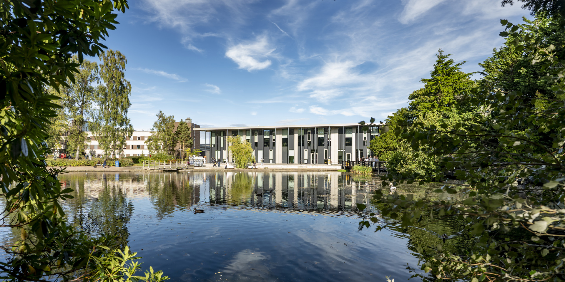 The modern GRID building reflecting in a serene Loch, surrounded by lush trees and clear blue skies.