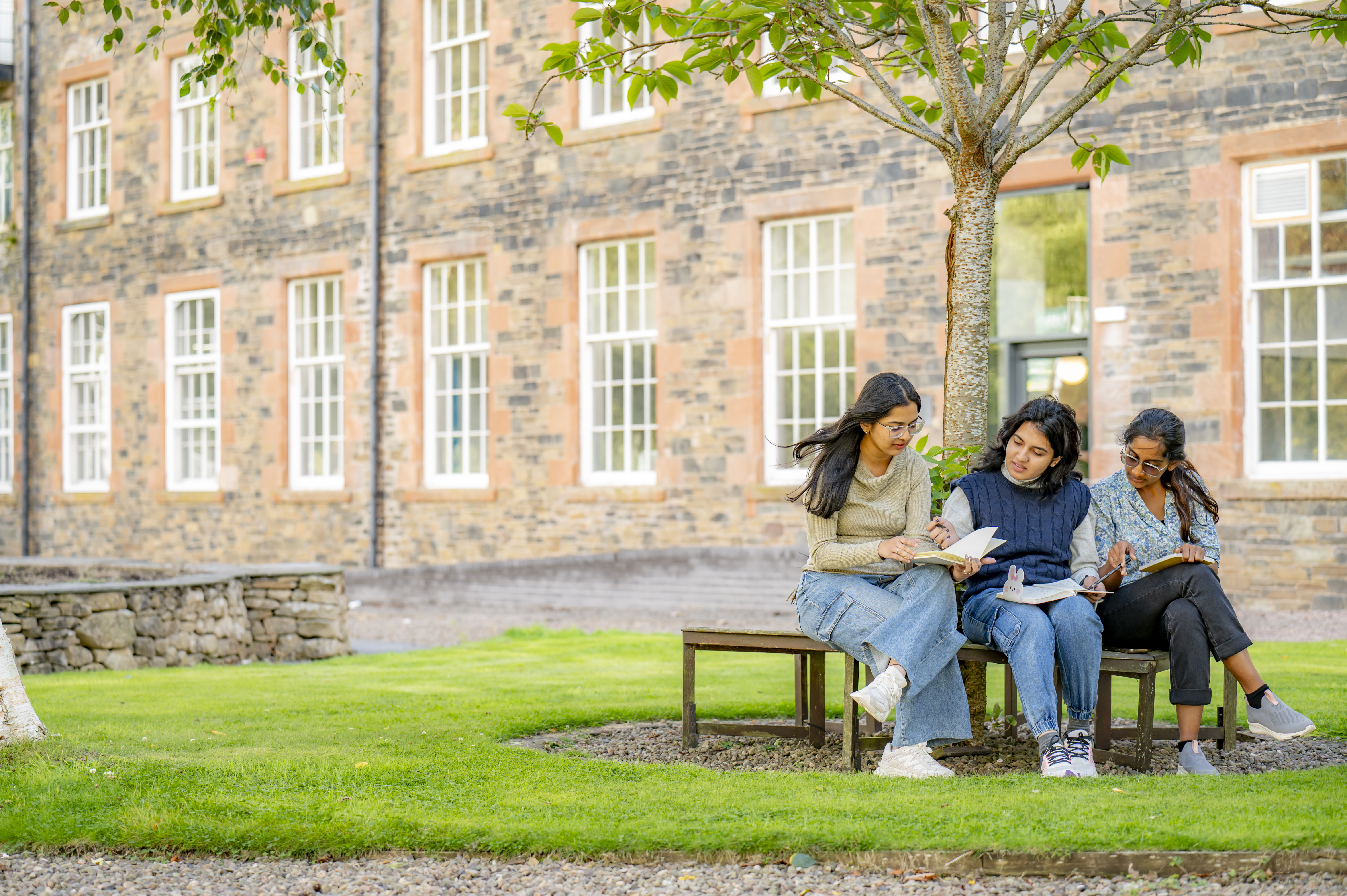 Three students sat on a bench underneath a tree on green grass, outside Heriot-Watt's Scottish Borders Campus building.