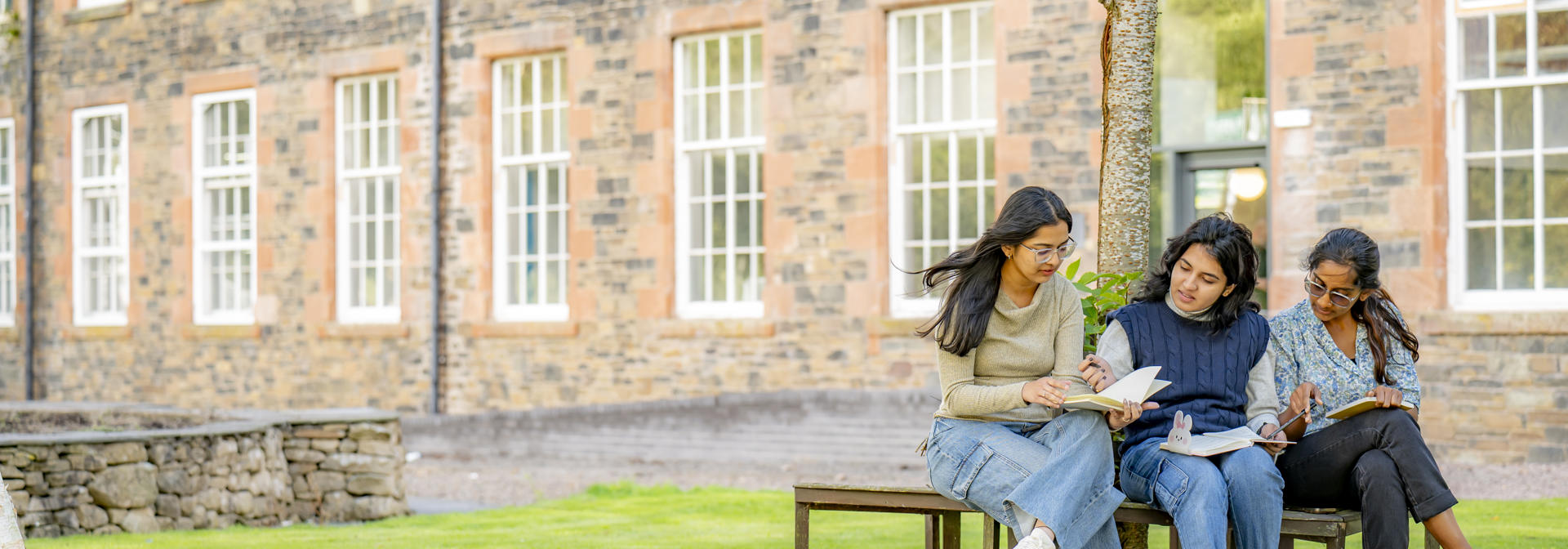 Three students sat on a bench underneath a tree on green grass, outside Heriot-Watt's Scottish Borders Campus building.