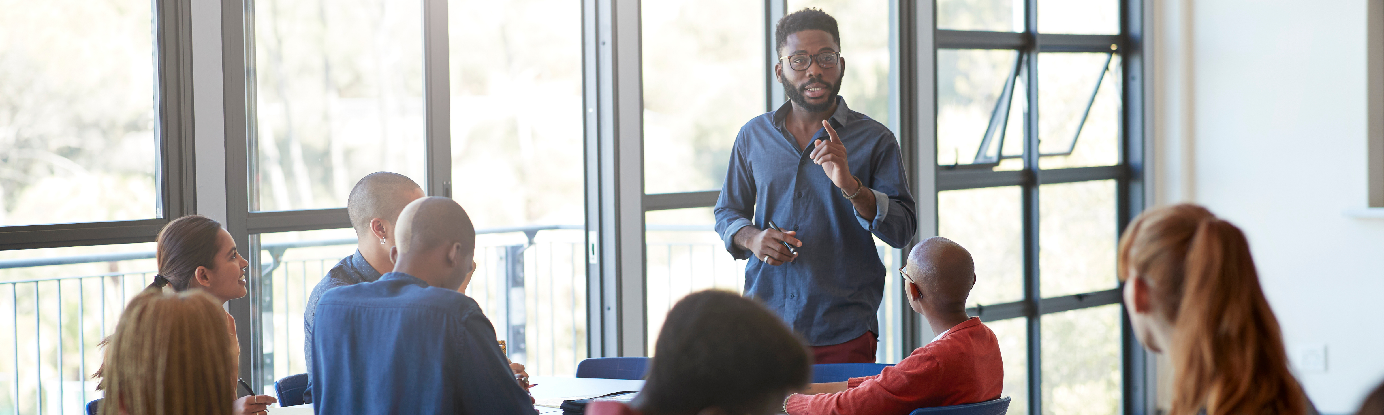 A man standing and leading a meeting consisting several people sitting down.