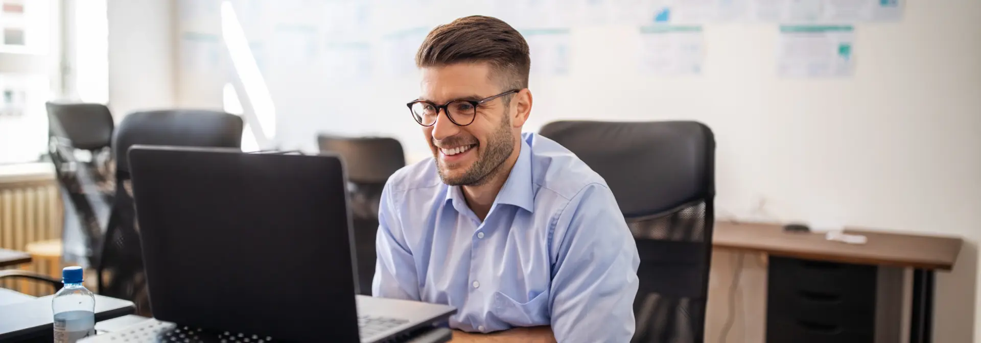 A man wearing glasses smiles as he sits working on a laptop.