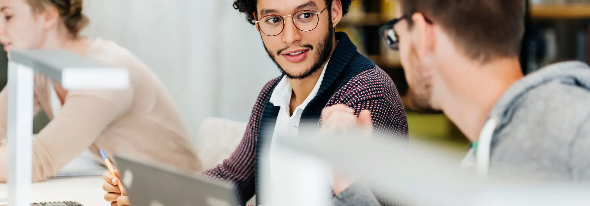 Two men wearing glasses in discussion while working on laptops in a library.