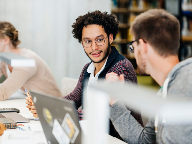 Two men wearing glasses in discussion while working on laptops in a library.