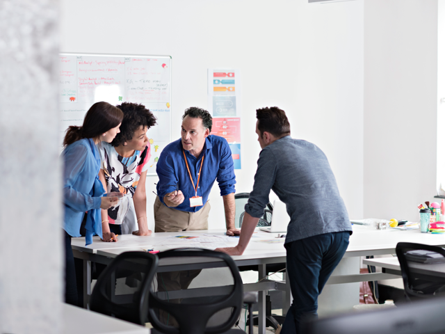 Four individuals lean over a table discussing a project intently.