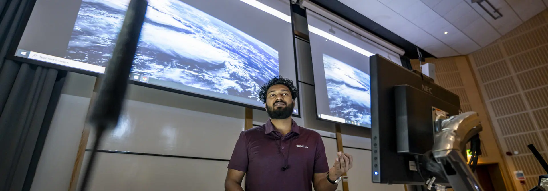 A man stands before a computer with a digital screen behind him, leading a lecture.