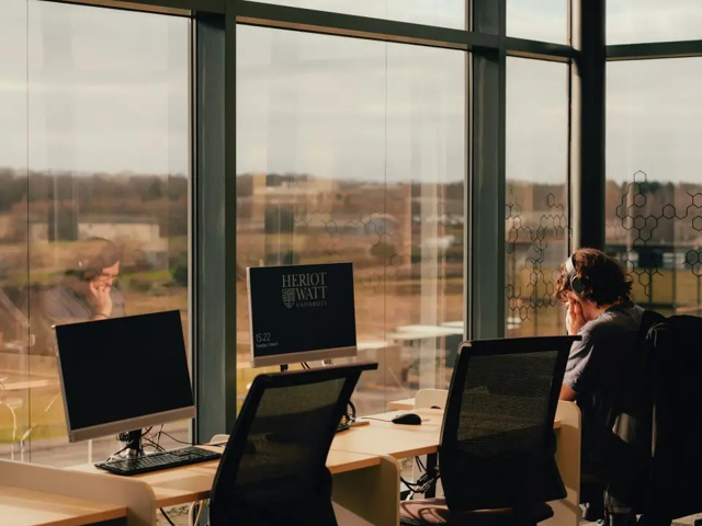 A man seated at a desk, working on a computer, with a large window providing natural light.