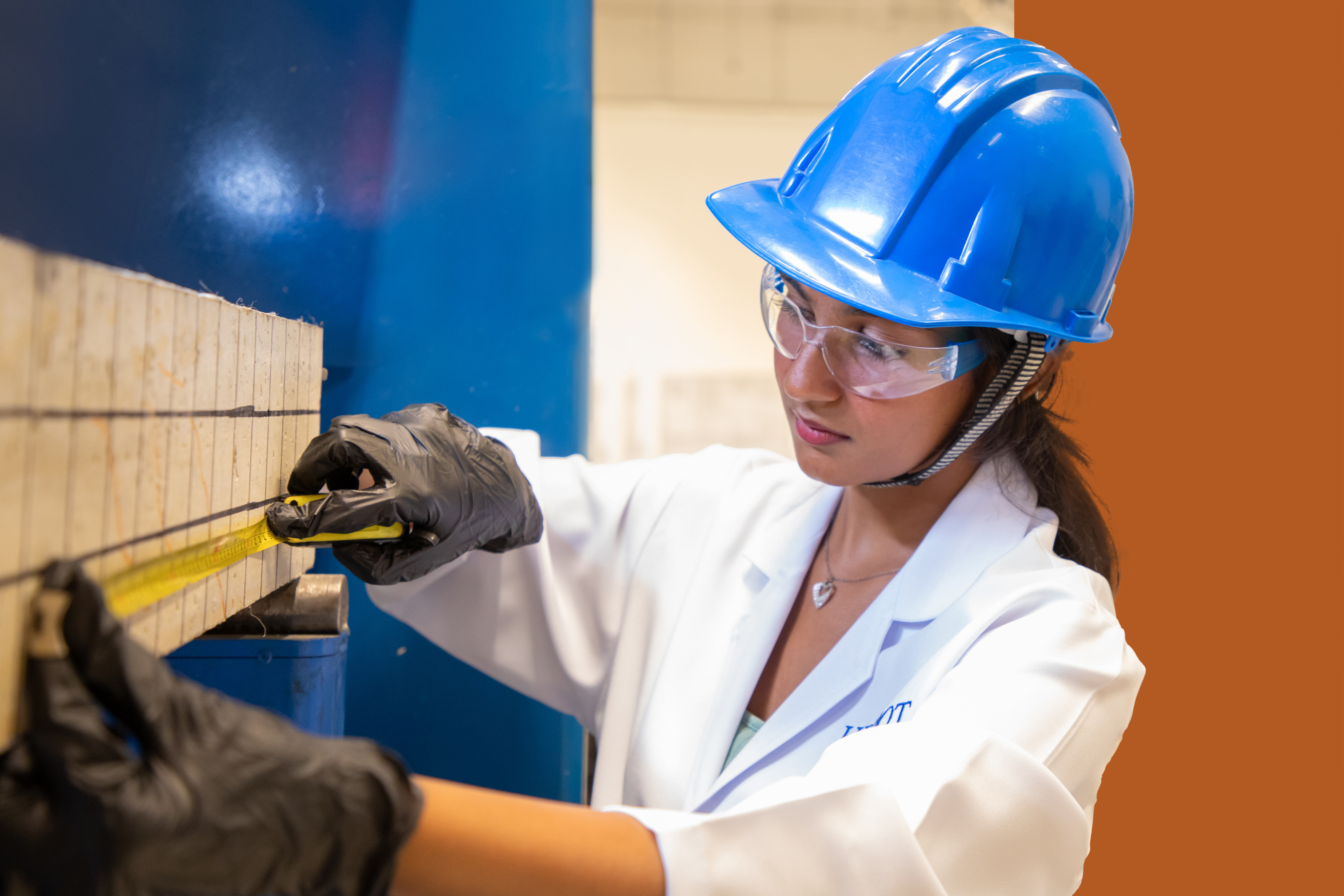 female student engineer measuring a piece of wood while wearing a white lab coat and blue safety helmet