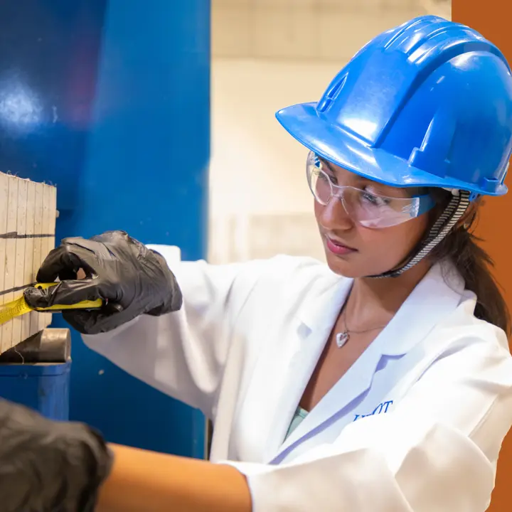 female student engineer measuring a piece of wood while wearing a white lab coat and blue safety helmet