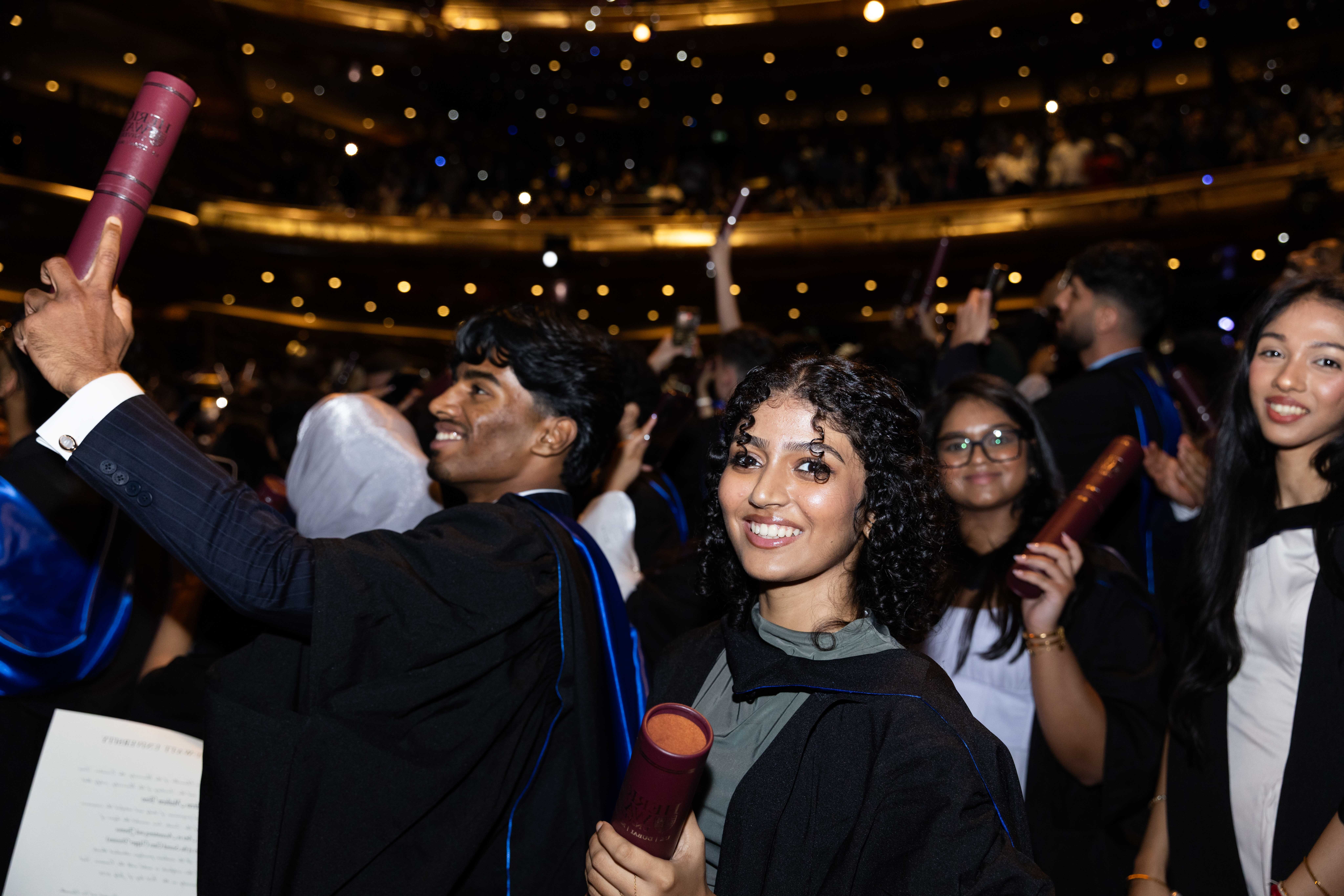 student wearing a graduation gown and holding a scroll looking at camera with other students cheering in the background 