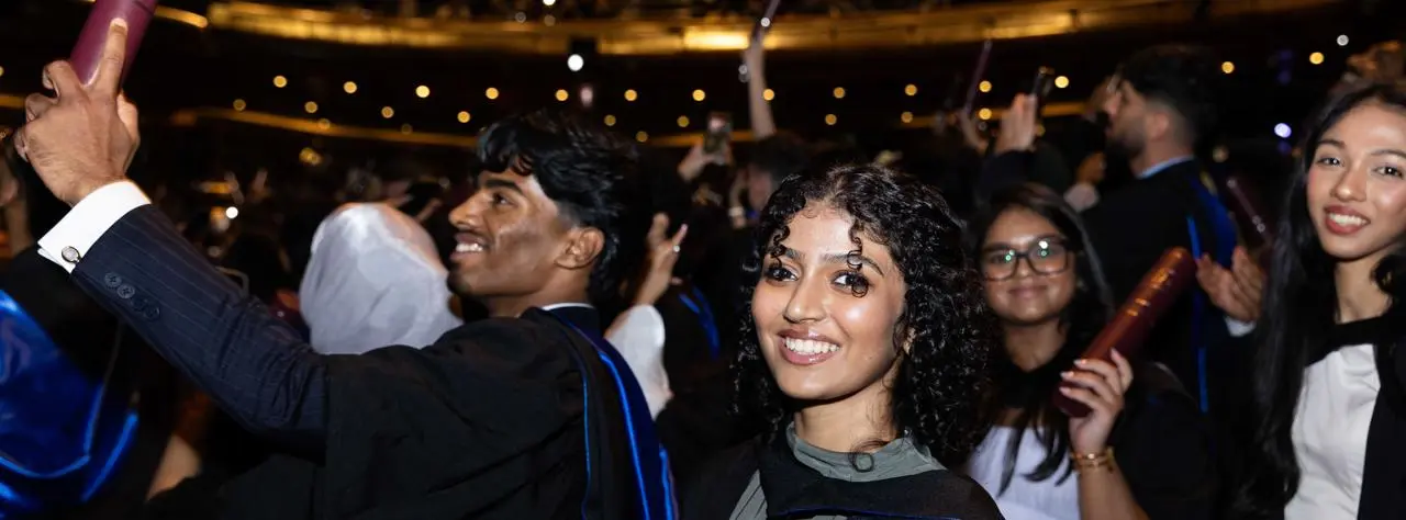 student wearing a graduation gown and holding a scroll looking at camera with other students cheering in the background