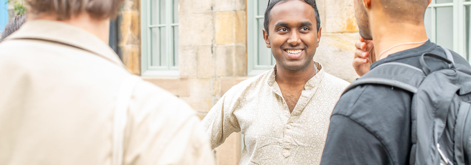 A smiling man speak with two individuals beside a stone wall.