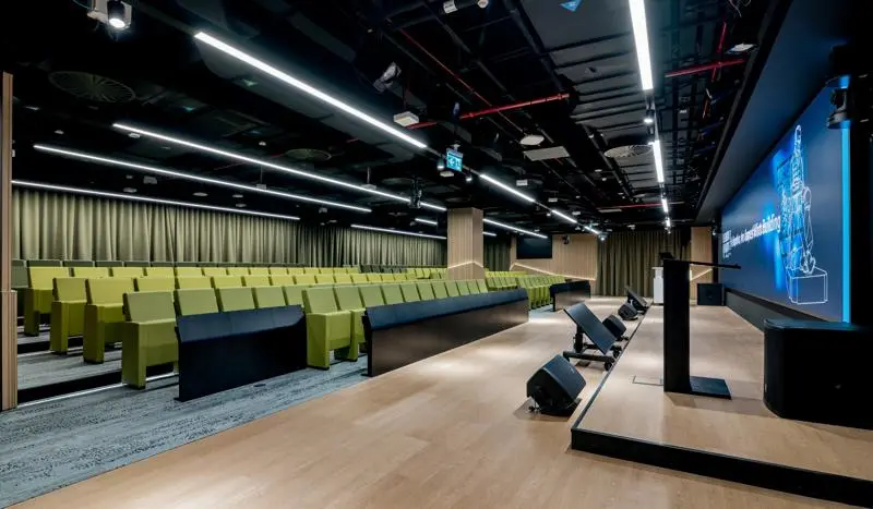 auditorium seating with green seats opposite a low stage infront of a video wall