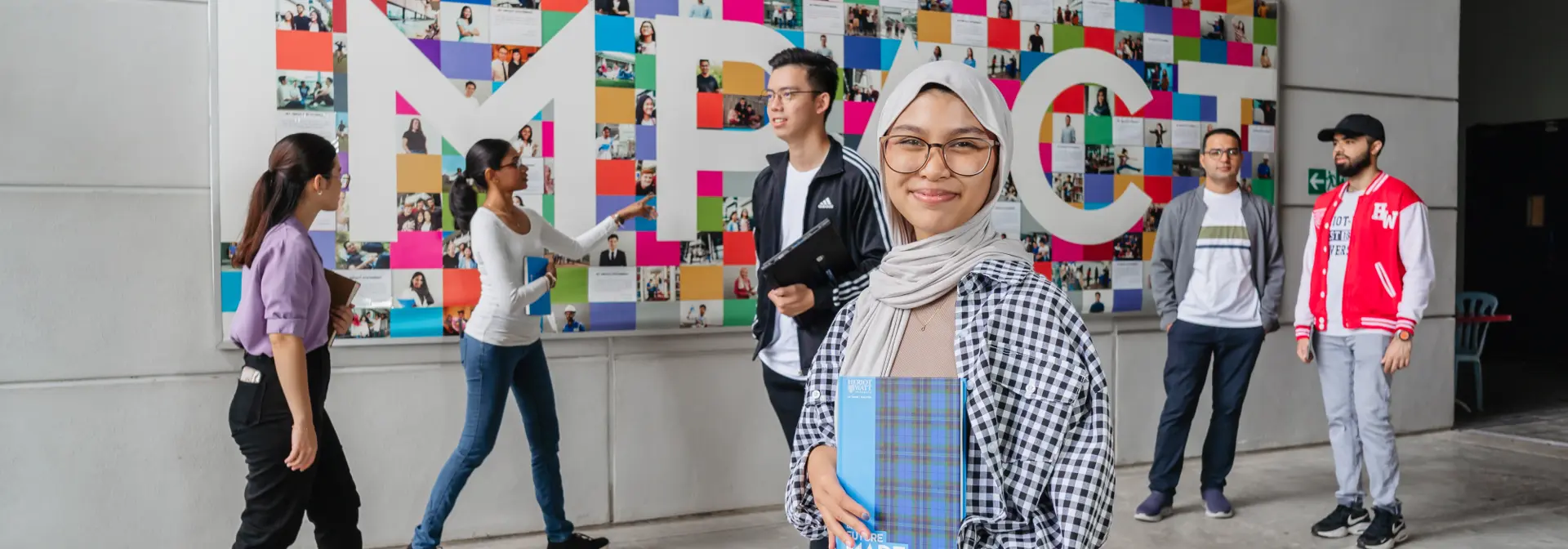 A woman stands in front of a colourful mural, people walking behind her as she smiles holding a book.