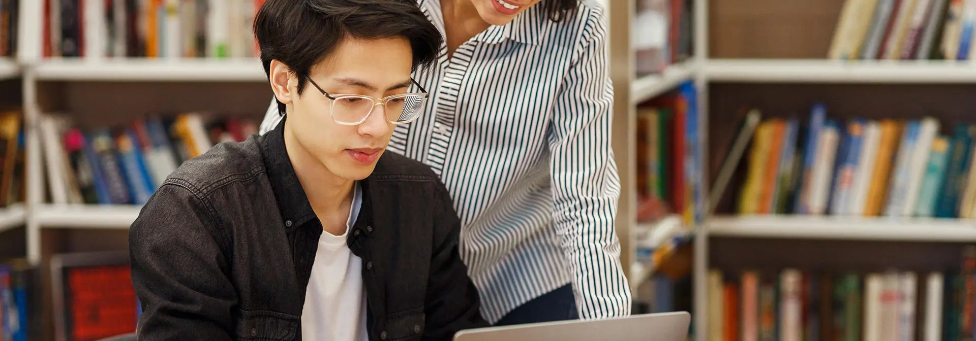 A man works on a laptop in a library with support of another behind him.