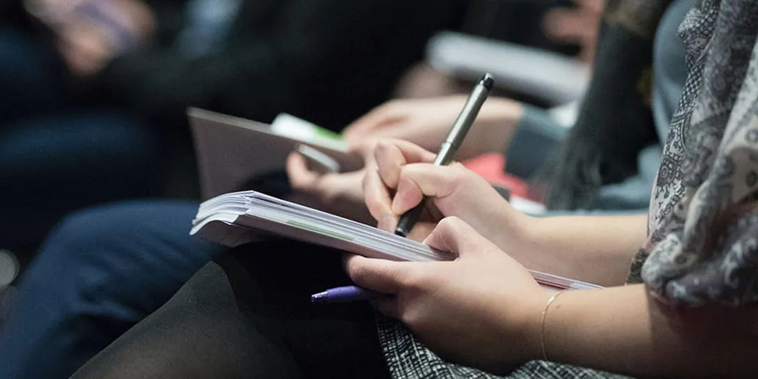 Person sitting at an event taking notes with a pen on a notepad.