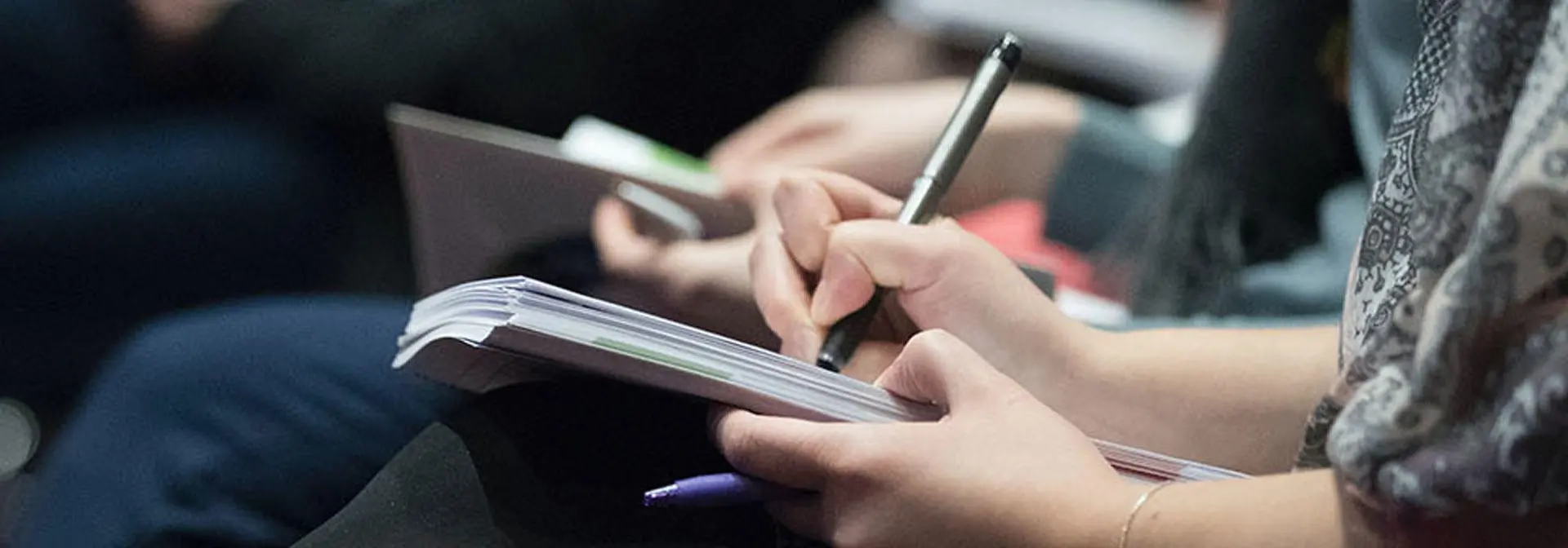 Person sitting at an event taking notes with a pen on a notepad.