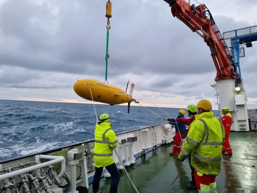 The image shows a group of individuals on the deck of a ship, wearing high-visibility clothing and hard hats. They are engaged in deploying or retrieving a large yellow torpedo-shaped object using a crane. 