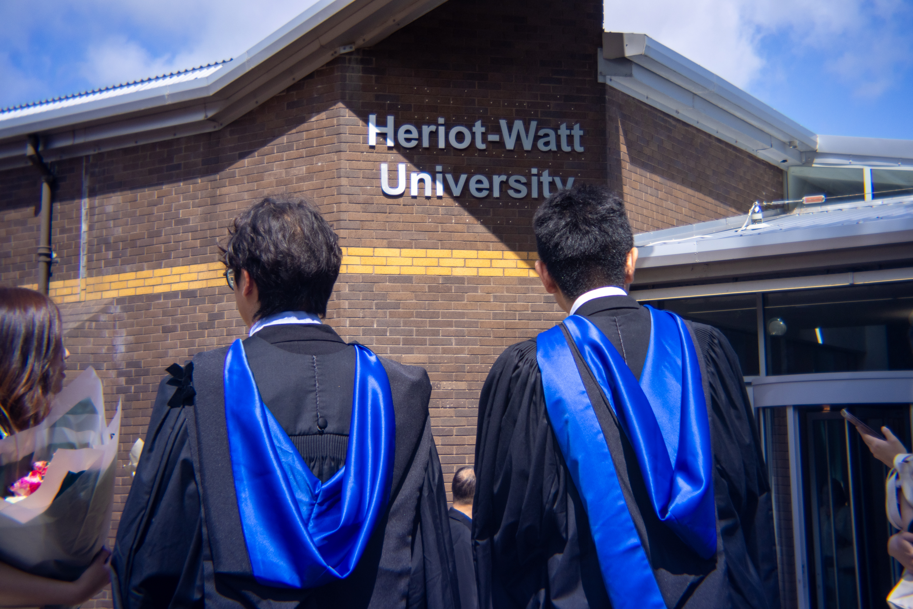 Shot shows two students standing in front of a sign that reads Heriot-Watt University.