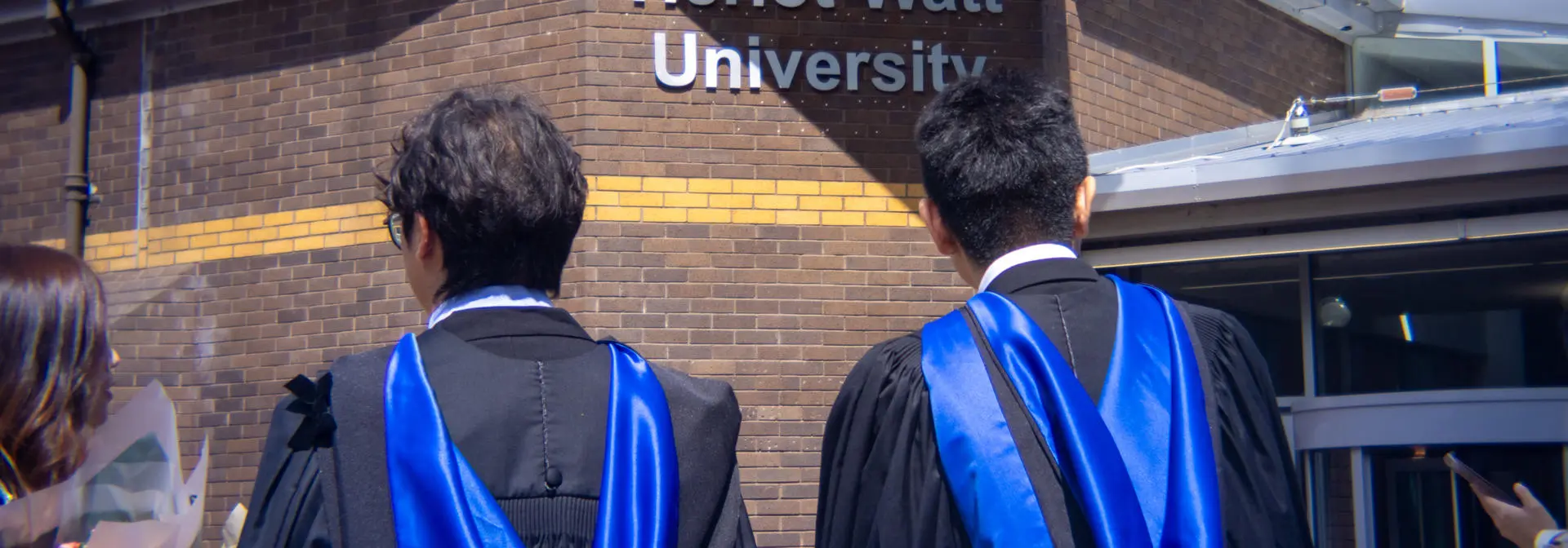 Shot shows two students standing in front of a sign that reads Heriot-Watt University.
