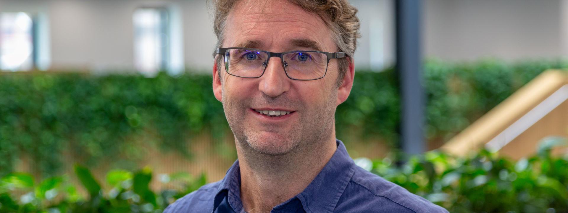 A man with wavy light brown hair, wearing glasses and a blue collared shirt, smiles warmly.