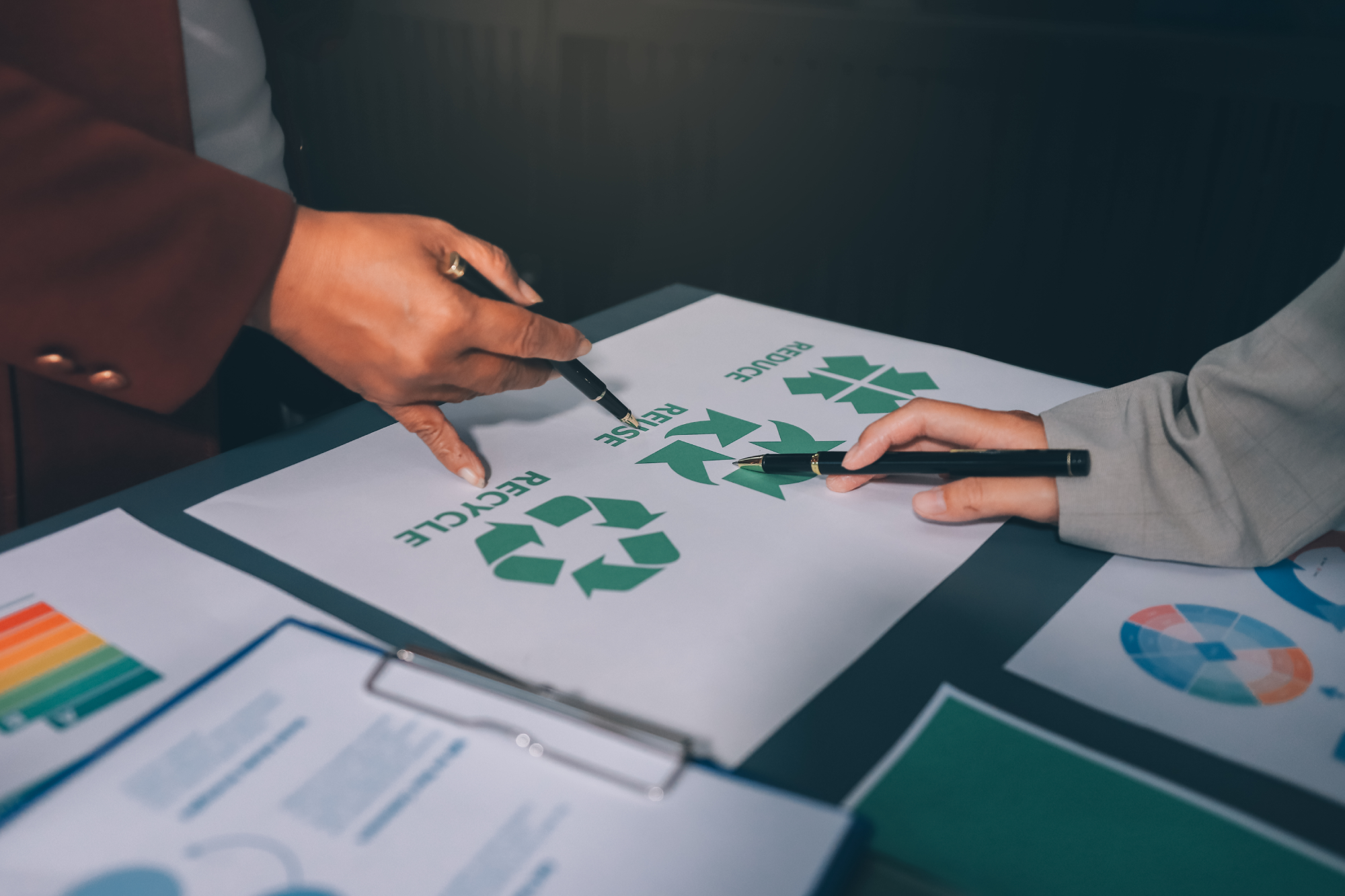 Two individuals in a room point with pens to a paper labelled reduce, reuse, recycle.
