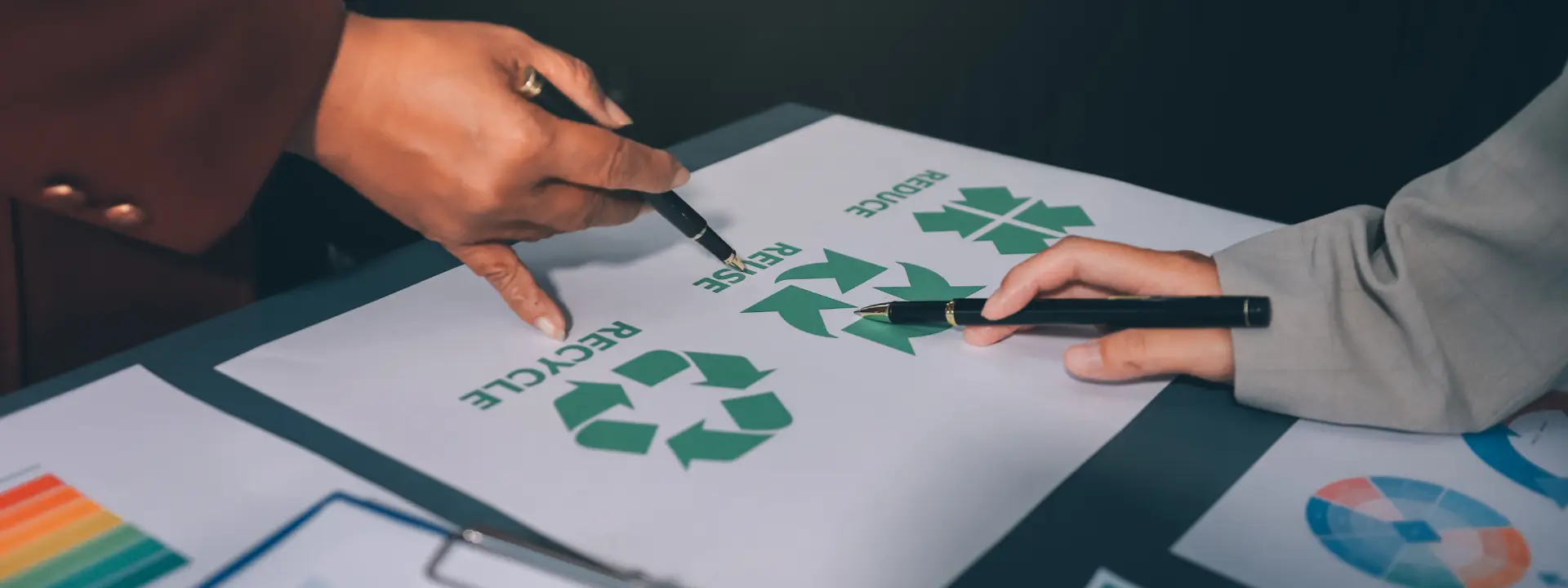 Two individuals in a room point with pens to a paper labelled reduce, reuse, recycle.