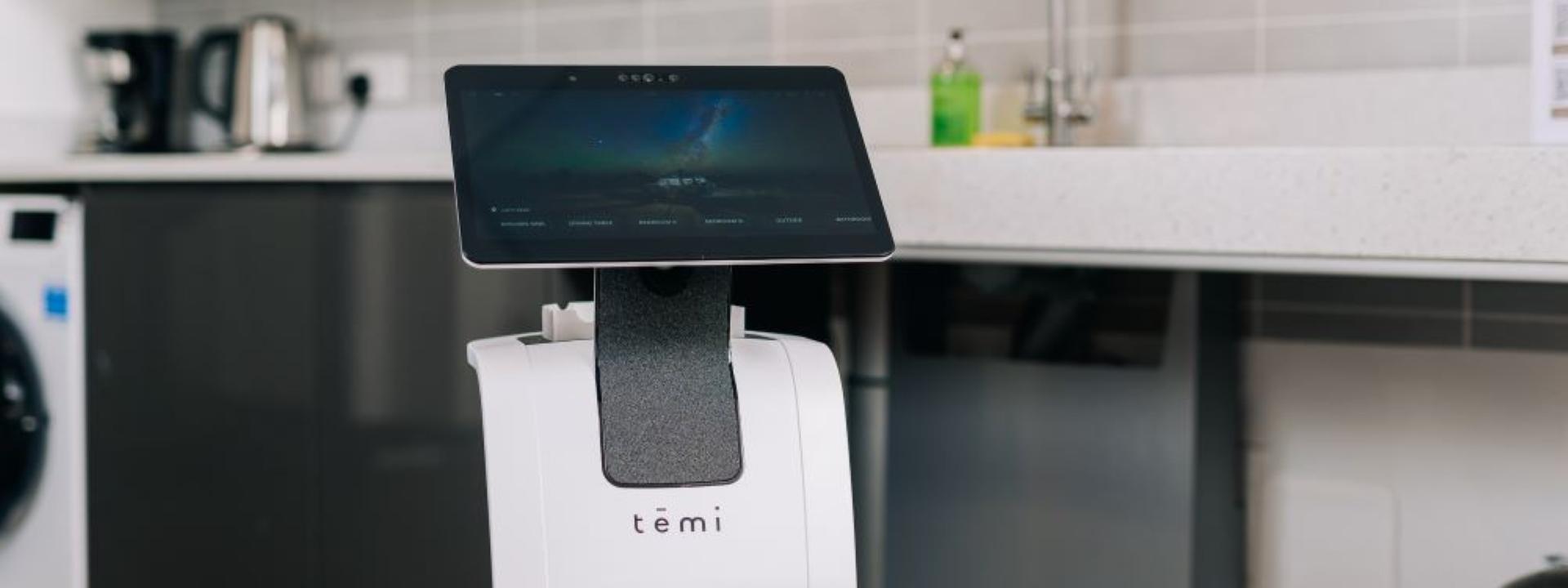 A white Temi service robot with a large touchscreen stands in the foreground of a modern kitchen
