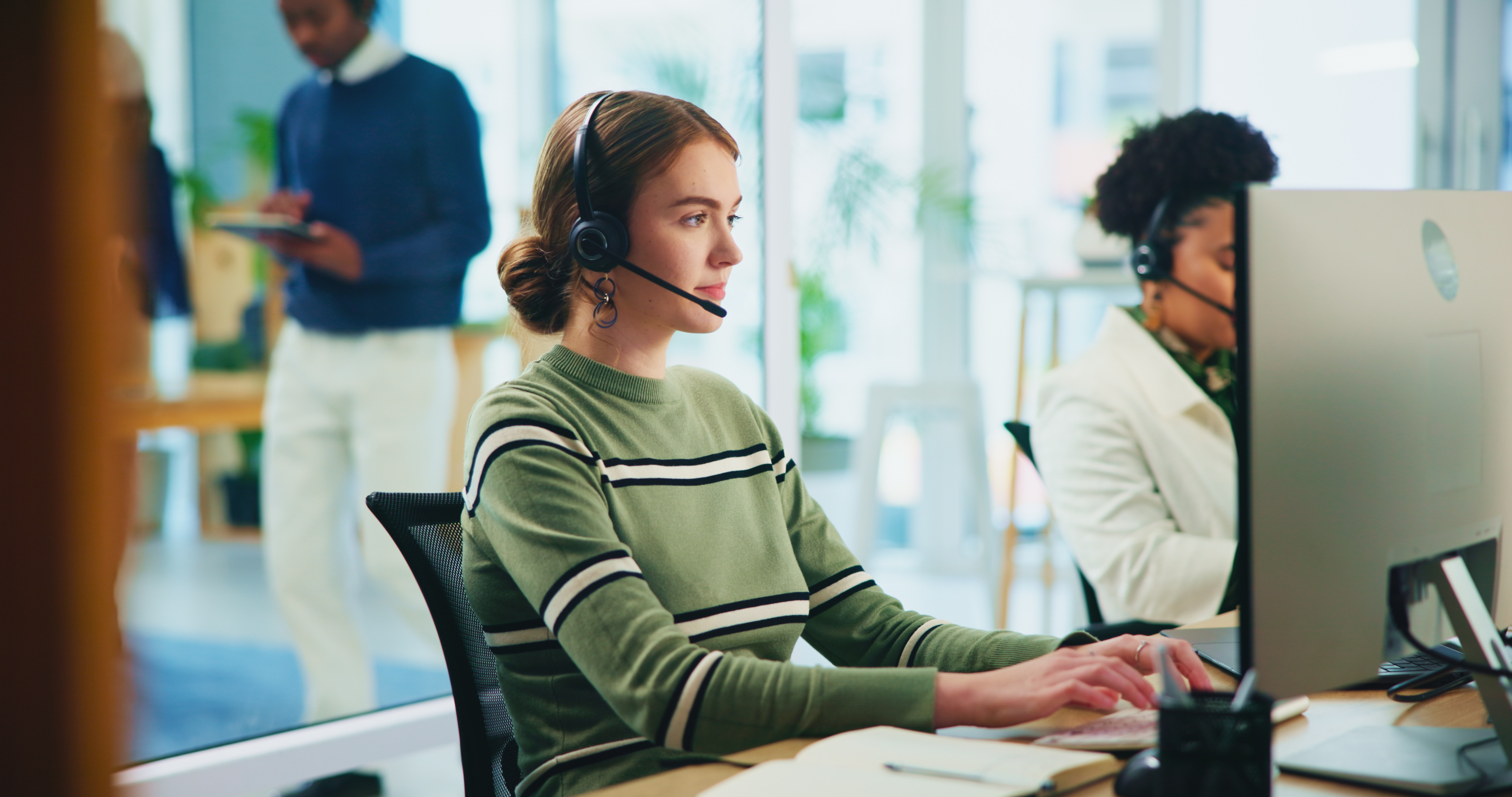A woman wearing a headset sits at a computer on the phone.