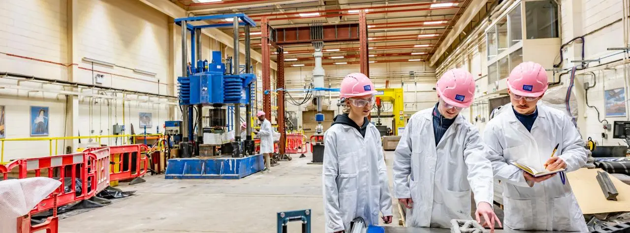 Three individuals wearing lab coats and safety helmets in warehouse.