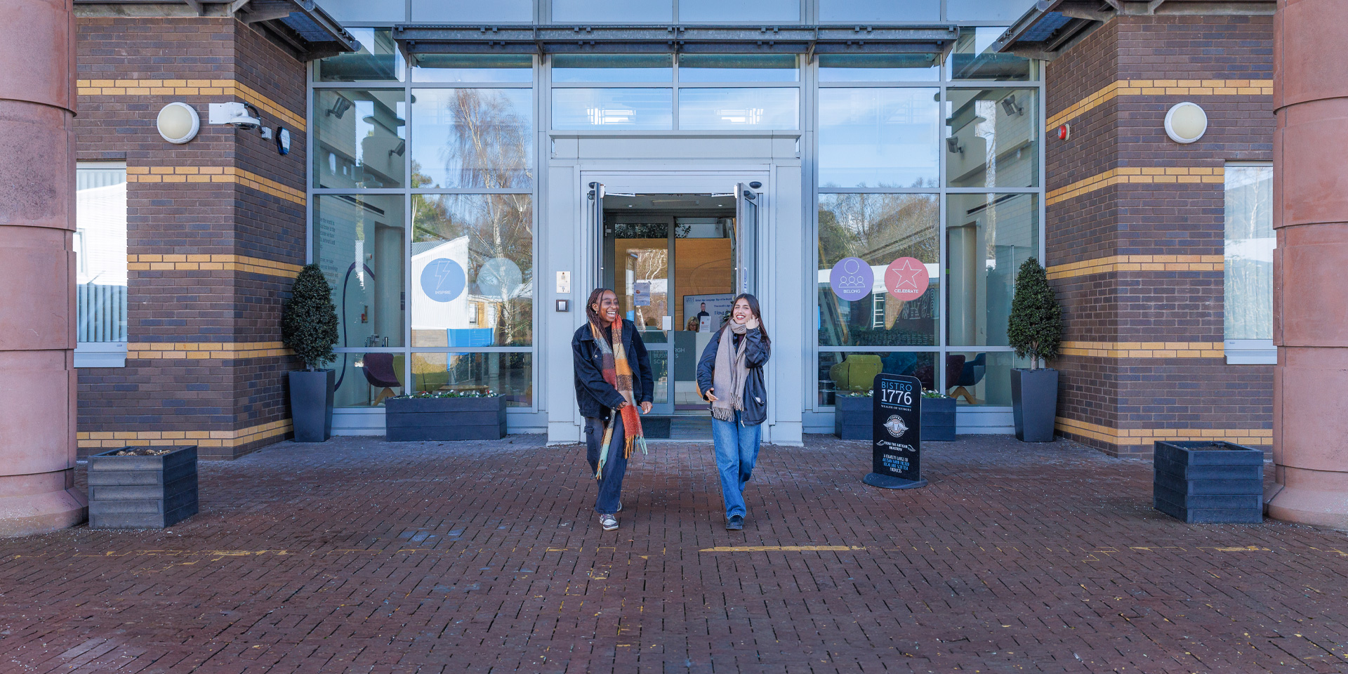 Two students outside the EBS building, with large glass doors and potted plants flanking the entrance.