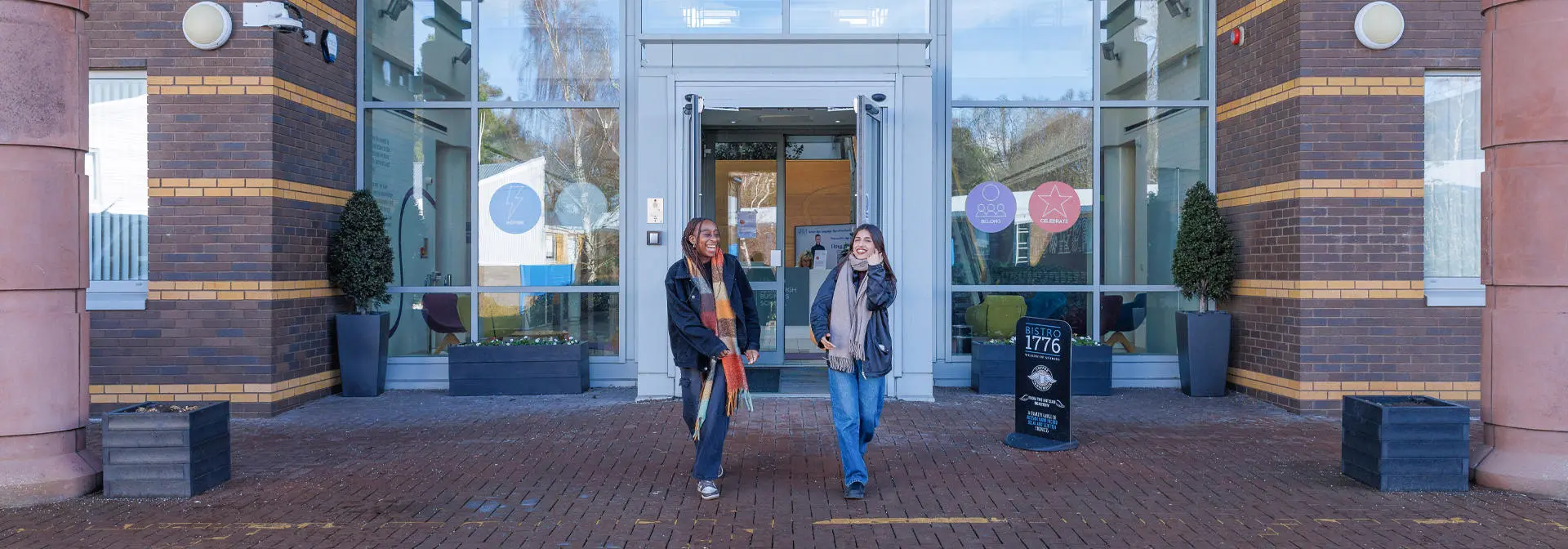 Two students outside the EBS building, with large glass doors and potted plants flanking the entrance.