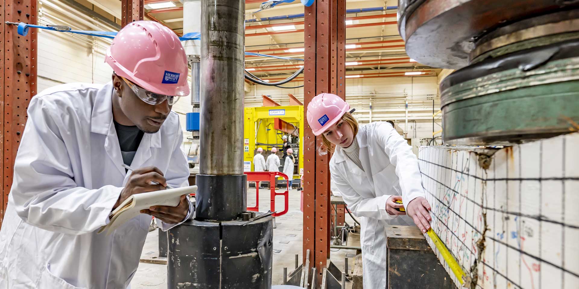 Two students in lab coats and hard hats, closely inspecting an item in a scientific setting.