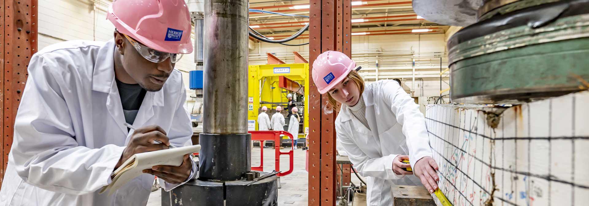 Two students in lab coats and hard hats, closely inspecting an item in a scientific setting.