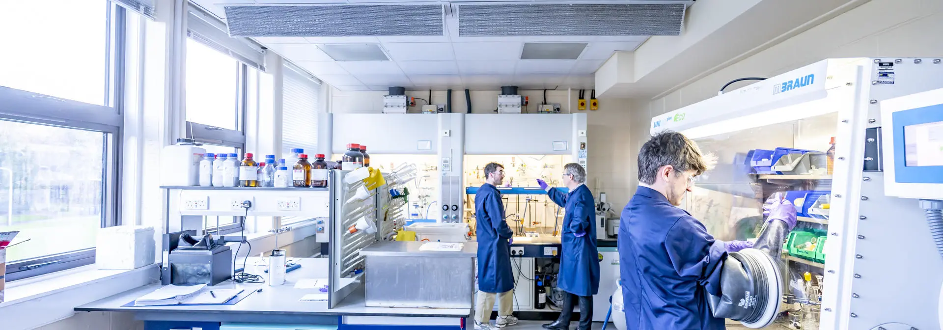 Two students in a chemistry lab, one using an isolation glove box and one talking with a member of staff