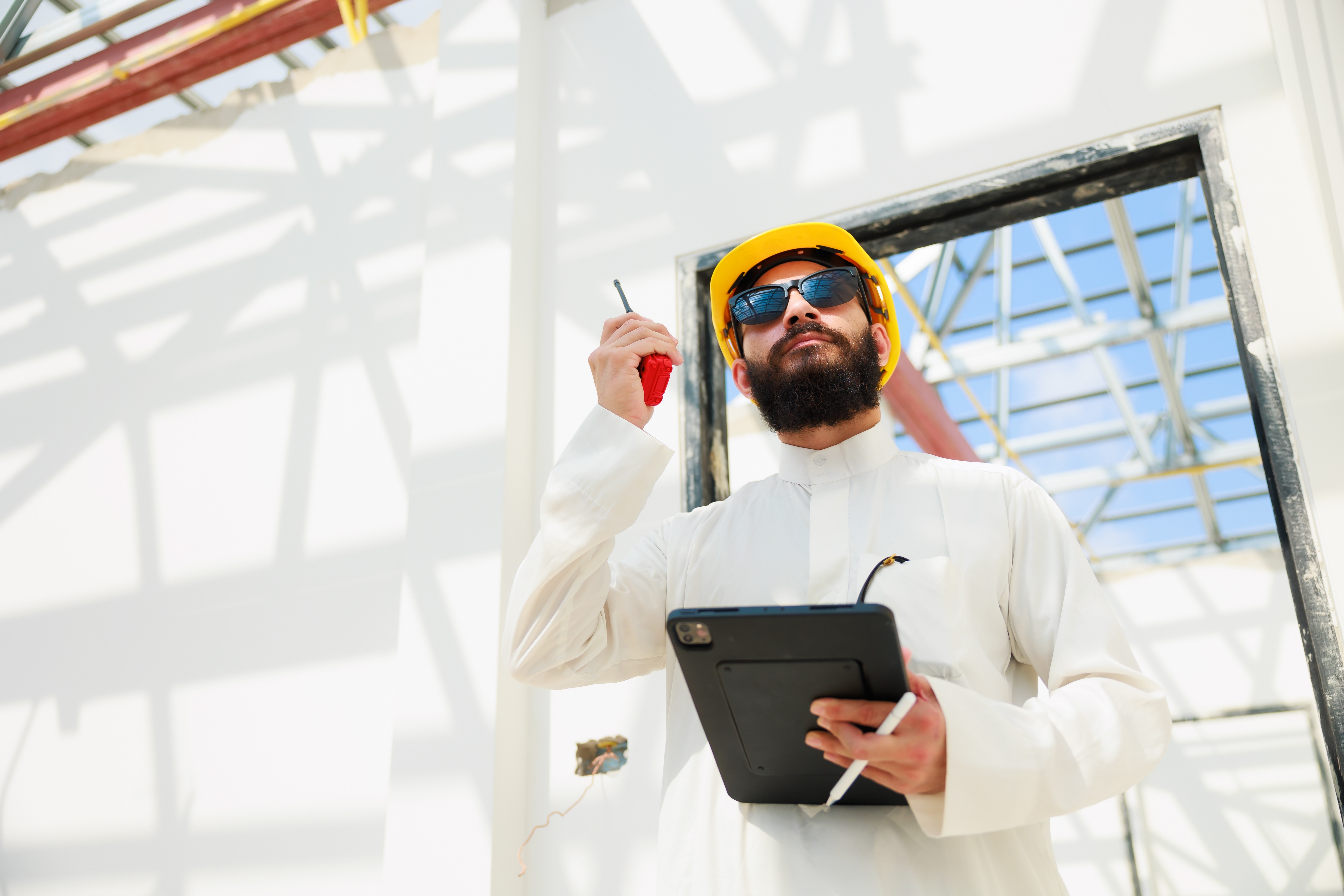 civil engineer in kandura and yellow hard hat inspecting a site talking into a walkie-talkie