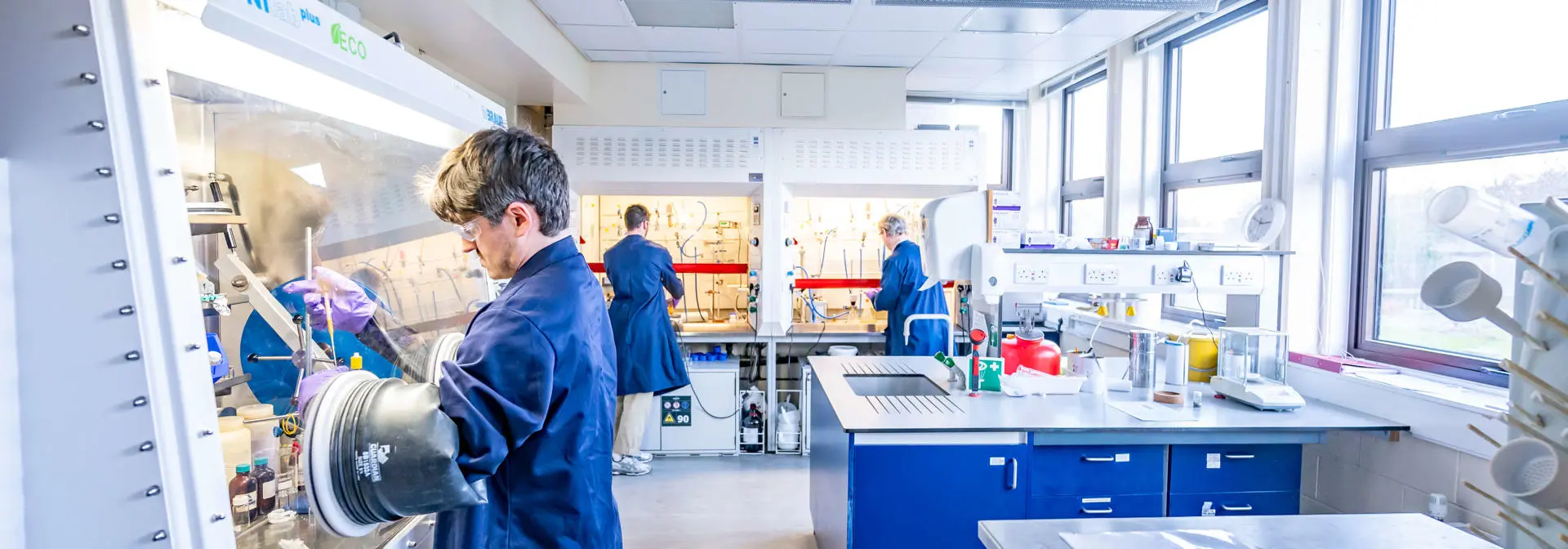 Two students in a chemistry lab, one using an isolation glove box and one talking with a member of staff