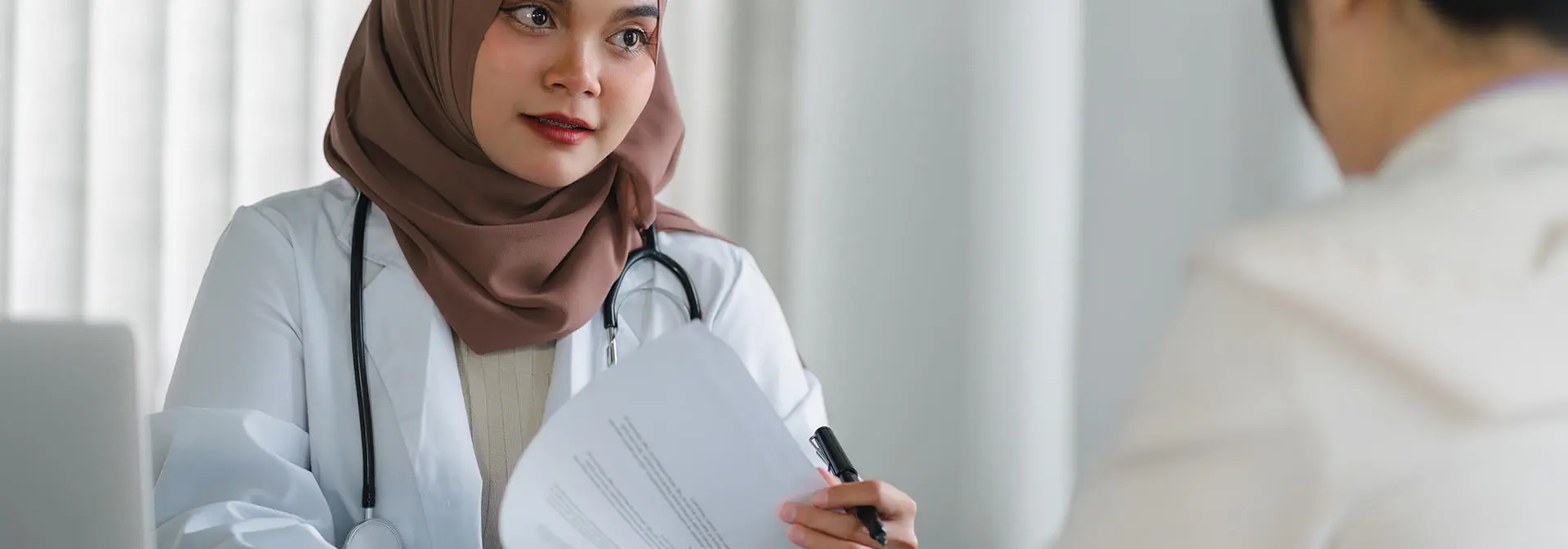 Doctor wearing a headscarf speaks at a desk to a person.