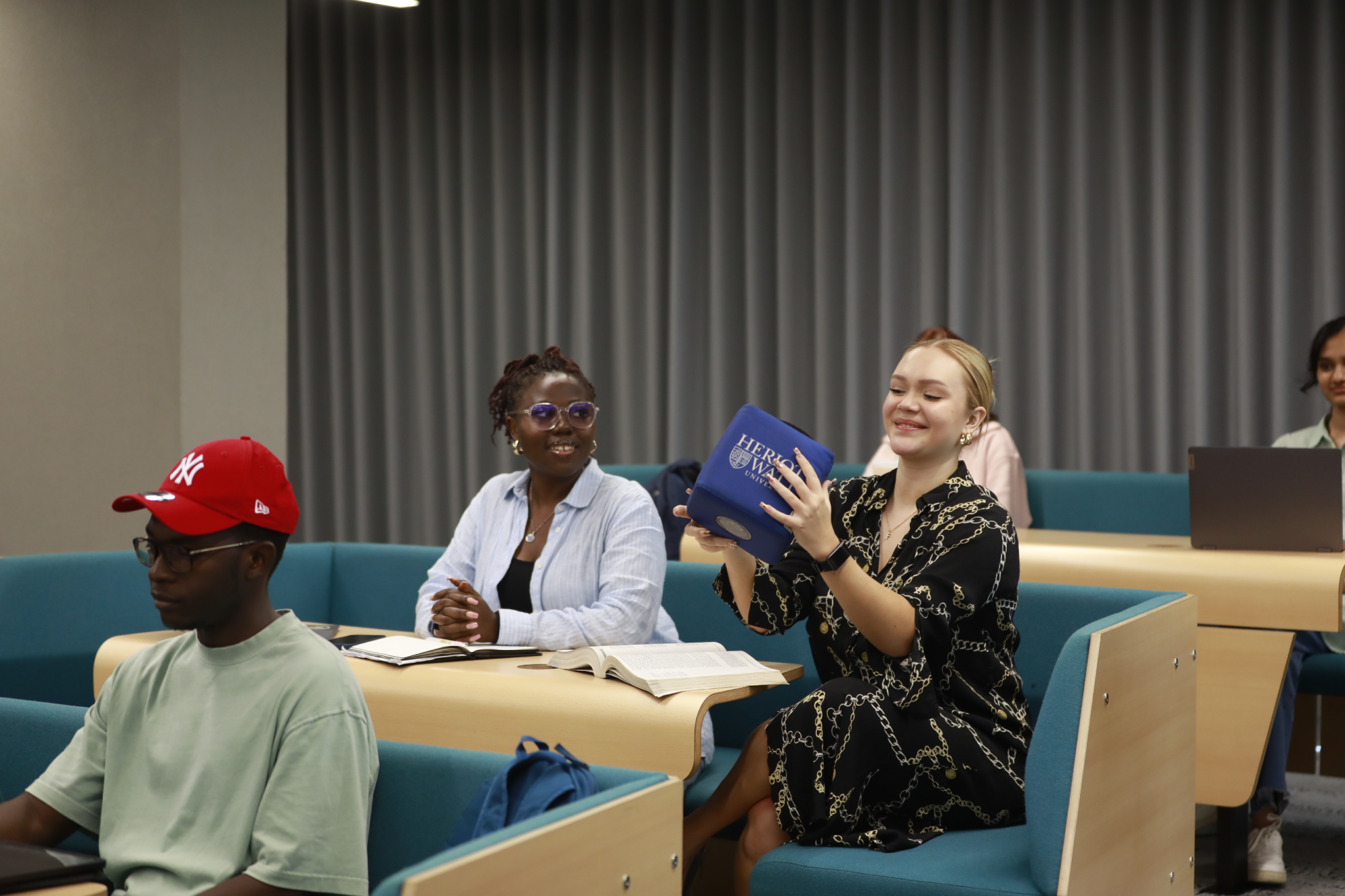 Three individuals sitting in a lecture theatre, smile as they pass around a cube.