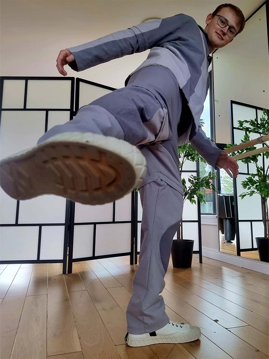 A man in a funky grey suit and white shoes karate kicking his foot towards the camera.