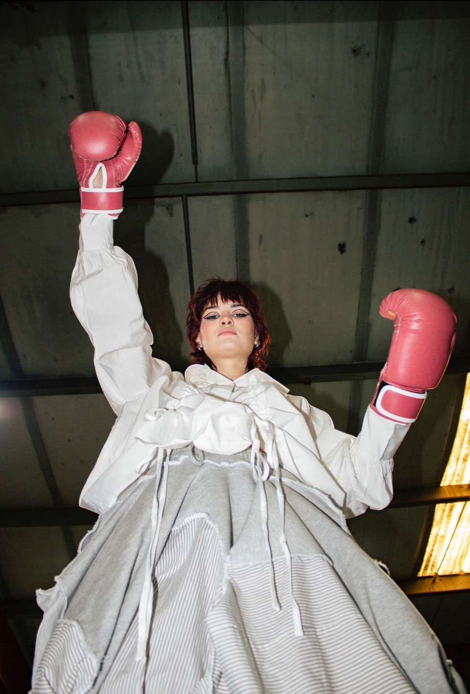 Young woman wearing white jacket, grey skirt and red boxing gloves holds her arms up while looking down at the viewer