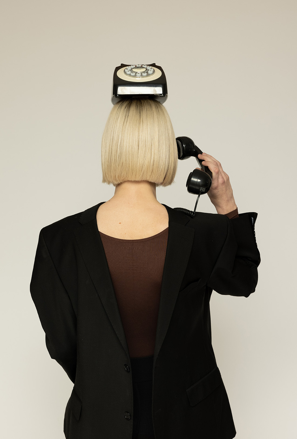 Rear view of a woman with black dress on backwards and old style bakelite telephone on top of her head, holds the receiver to the back of her head