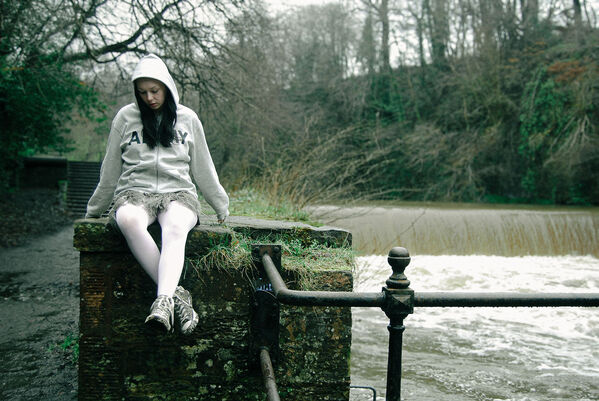 Young woman weaing white hoodie, skirt with feathered frontpiece, white tights and camouflage trainers sits on a wall above a weir on an overcast day
